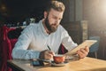 Young bearded businessman sits in cafe, home at table, uses tablet computer and writes in notebook. Royalty Free Stock Photo