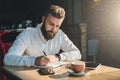 Young bearded businessman sits in cafe, home at table and writes in notebook. On table tablet computer, smartphone.