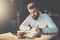 Young bearded businessman sits in cafe at table and writes in notebook.On table tablet computer, smartphone.Man is