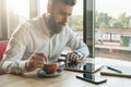 Young bearded businessman sits in office at table,uses tablet computer, drinks coffee.On desk is notebook,smartphone. Royalty Free Stock Photo