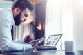 Young bearded businessman sitting at desk in front of laptop with graphis, charts, diagrams on screen Royalty Free Stock Photo