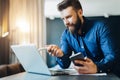 Young bearded businessman is sitting in front of computer, holding smartphone and showing pen on laptop screen. Royalty Free Stock Photo