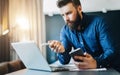 Young bearded businessman is sitting in front of computer, holding smartphone and showing pen on laptop screen. Royalty Free Stock Photo