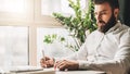 Young bearded businessman is sitting at table in office in front of laptop near window and writing in notebook. Royalty Free Stock Photo