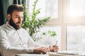 Young bearded businessman is sitting at table in office in front of laptop near window and writing in notebook. Royalty Free Stock Photo