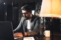 Young bearded businessman sitting at wooden table with lamp in loft office at night. Project manager working process and typing te