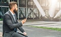 Young bearded businessman in suit and tie sitting in park on bench, holding closed laptop and using smartphone Royalty Free Stock Photo