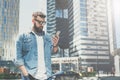 Young bearded businessman in sunglasses stands on city street and uses smartphone. Man looking on screen of phone. Royalty Free Stock Photo