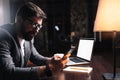 Young bearded businessman using phone while sitting by the wooden table in modern office at night. People working mobile devices