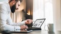 Young bearded businessman in white shirt is sitting at table in front of computer, pointing with pen on graphs, charts Royalty Free Stock Photo