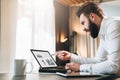 Young bearded businessman in white shirt is sitting at table in front of computer, pointing with pen on graphs, charts Royalty Free Stock Photo