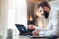 Young bearded businessman in white shirt is sitting at table in front of computer,pointing with pen on laptop screen.