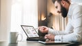 Young bearded businessman in white shirt is sitting at table in front of computer, showing pen on laptop screen Royalty Free Stock Photo