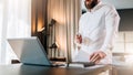 Young bearded businessman in white shirt is standing near desk in front of laptop, holding documents. Freelancer working Royalty Free Stock Photo