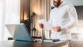 Young bearded businessman in white shirt is standing near desk in front of laptop, reading documents. Freelancer working Royalty Free Stock Photo