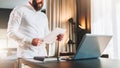 Young bearded businessman in white shirt is standing near desk in front of laptop, reading documents. Freelancer working Royalty Free Stock Photo