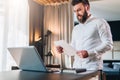 Young bearded businessman in white shirt is standing near desk in front of laptop, reading documents. Freelancer working Royalty Free Stock Photo