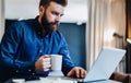 Young bearded businessman working on computer at table, drinking coffee.Man analyzes information, develops business plan