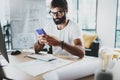 Young bearded graphic designer wearing eye glasses and working at modern loft studio-office.Man using smartphone.Blurred Royalty Free Stock Photo