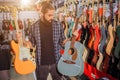 Young bearded guy stand and hold electric and acoustic guitars. He looks at blue one. Young hipster in alone in room