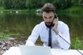 Young, bearded guy on worktable in summer, using laptop, talking on phone. man working, studying or surfing web in park Royalty Free Stock Photo