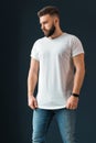 Young bearded handsome hipster man, dressed in white T-shirt with short sleeves and jeans, stands indoors