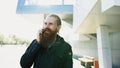 Young bearded hipster man concentrated talking on phone on citystreet, conversation near office building Royalty Free Stock Photo