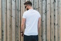 Young bearded hipster man dressed in white t-shirt and sunglasses is stands outdoor against wood wall. Mock up.