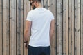 Young bearded hipster man dressed in white t-shirt and sunglasses is stands outdoor against wood wall. Mock up.