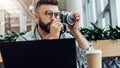 Young bearded man sits in coffee shop at table in front of laptop and takes instant photo on camera. Royalty Free Stock Photo