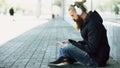 Young bearded hipster man with headphones sitting on road and using smartphone for listen to music and internet surfing