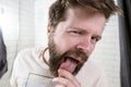 A young, bearded man alarmed looks at his tongue in the reflection of a mirror in the bathroom