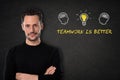 Young bearded man with crossed arms, heads with brains, lightbulb-idea and text `Teamwork is better` on a blackboard background. Royalty Free Stock Photo