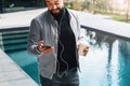 Young bearded man listens to music through headphones on smartphone and drinks coffee while standing outdoors. Lifestyle Royalty Free Stock Photo