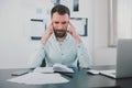 Young bearded man looking miserable suffering extreme headache at work while studying business project in his modern office, holds