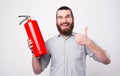 A young bearded man is looking and smiling at the camera holding a fire extinguisher and a thumg up near a white wall Royalty Free Stock Photo