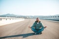 Young bearded man is meditating while sitting on the road or highway in easy lotus pose