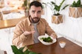 Young bearded man seems to be unhappy with quality and taste of his salad during lunch at the cafe, complaints and suggestions