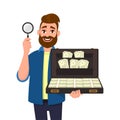 Young bearded man showing/holding magnifying glass and brief case full of money/cash/currency bank notes. Search, find, discovery.