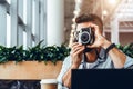 Young bearded man sits in coffee shop at table in front of laptop and takes instant photo on camera. Royalty Free Stock Photo