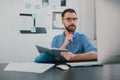Young bearded man sits in office holding pen near face while working on business project looks satisfied, takes notes to Royalty Free Stock Photo