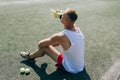 Young bearded man sitting on basketball court outdoors with ball holding bottle drinking electrolytes drink.