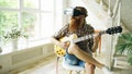 Young bearded man sitting on chair learning to play guitar using VR 360 headset and feels him guitarist at concert at