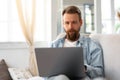 Young bearded man sitting on sofa at home using laptop computer Royalty Free Stock Photo