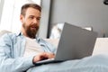 Young bearded man sitting on sofa at home using laptop computer Royalty Free Stock Photo