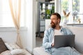 Young bearded man sitting on sofa at home using laptop computer Royalty Free Stock Photo