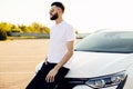 Young bearded man standing near a car outdoors on a summer day, a man in a white shirt stands next to a white car Royalty Free Stock Photo