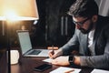 Young bearded man wearing glasses, smartwatch and office wear using modern laptop at night loft office. Brainstorming process. Bus