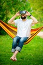 Young bearded man wearing virtual reality goggles relaxing in a garden hammock. Lifestyle VR fun Royalty Free Stock Photo