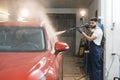 Young bearded man worker washing red crossover car under high pressure water in car wash service. Royalty Free Stock Photo